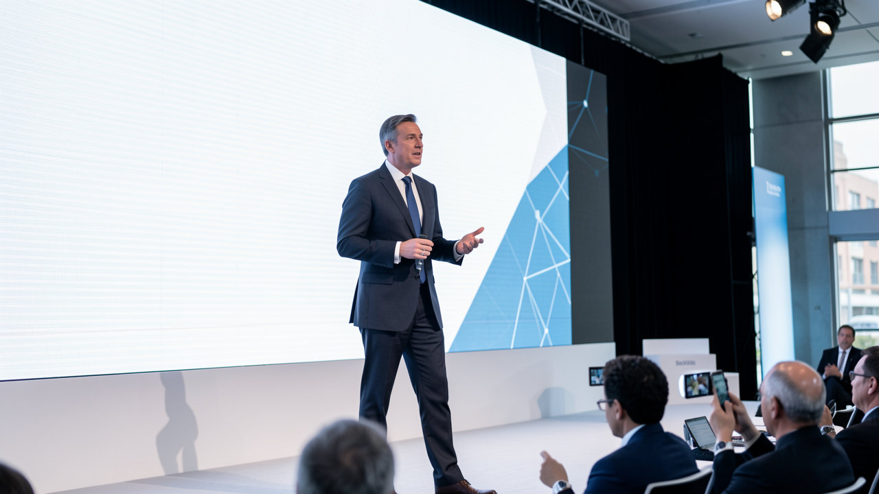 A business executive speaking on stage at a modern travel technology conference with a large screen behind him.