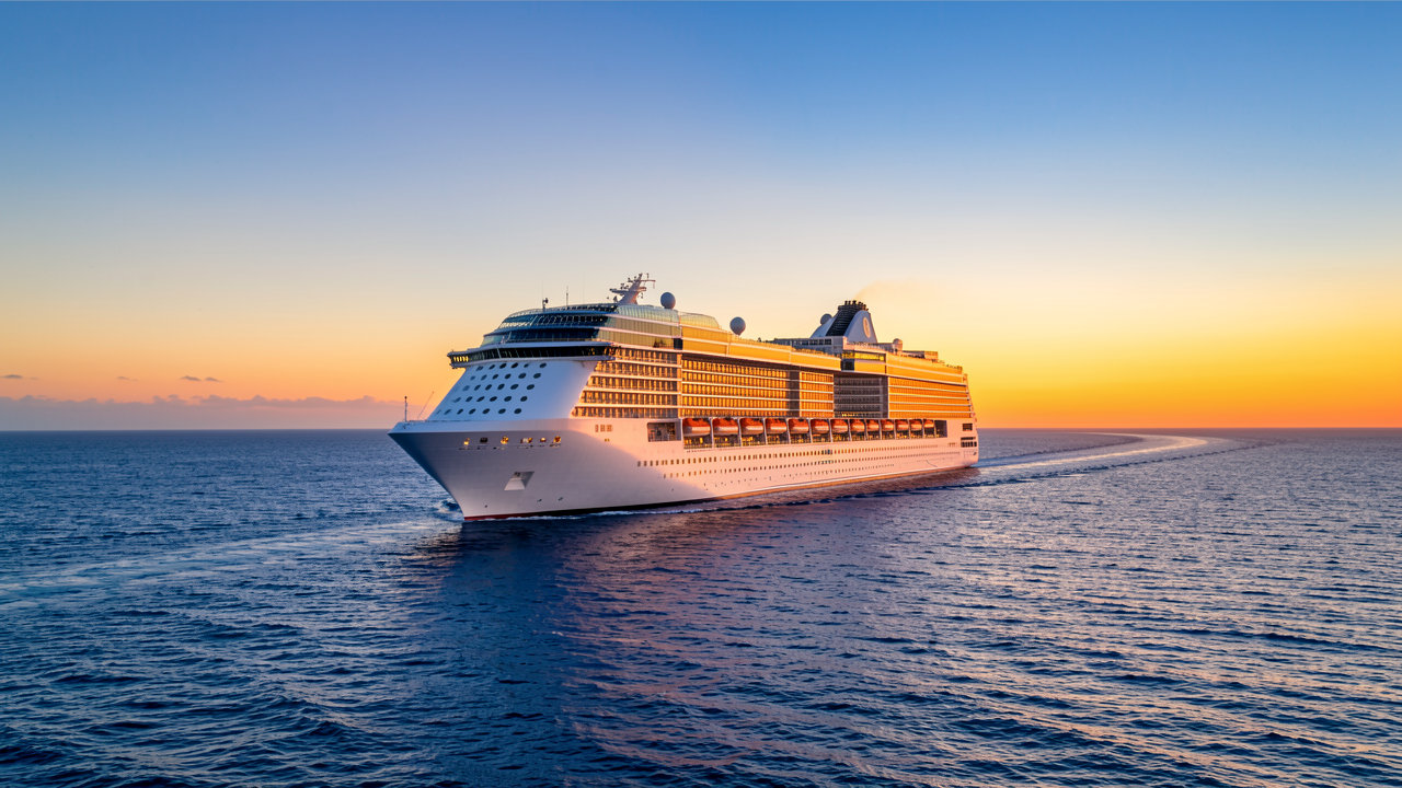 A modern cruise ship sailing on blue ocean waters under a clear sky with a vibrant sunset.