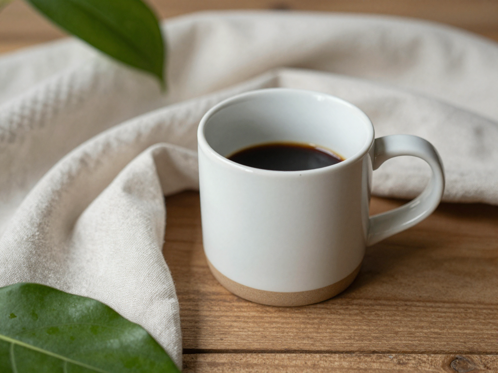 나무 테이블 위 머그잔과 초록 잎사귀, 부드러운 린넨 천이 어우러진 따뜻하고 차분한 분위기의 실내 사진.